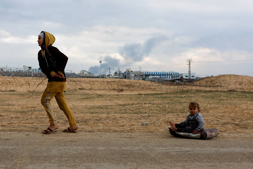 Рафах, сектор Газа. Женщина тащит ребенка, сидящего на сумке, на фоне дыма от израильского наступления