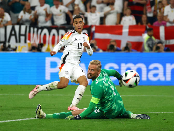 Полузащитник сборной Германии Джамал Мусиала отправляет мяч в ворота датского голкипера Каспера Шмейхеля (справа)