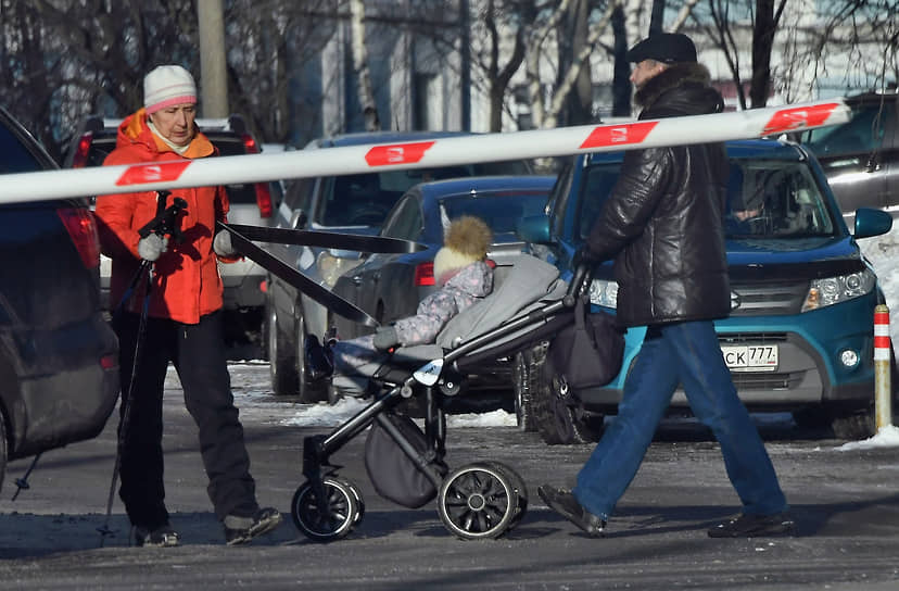 Женщина с лыжами и мужчина с ребенком в коляске перед шлагбаумом во дворе жилого дома